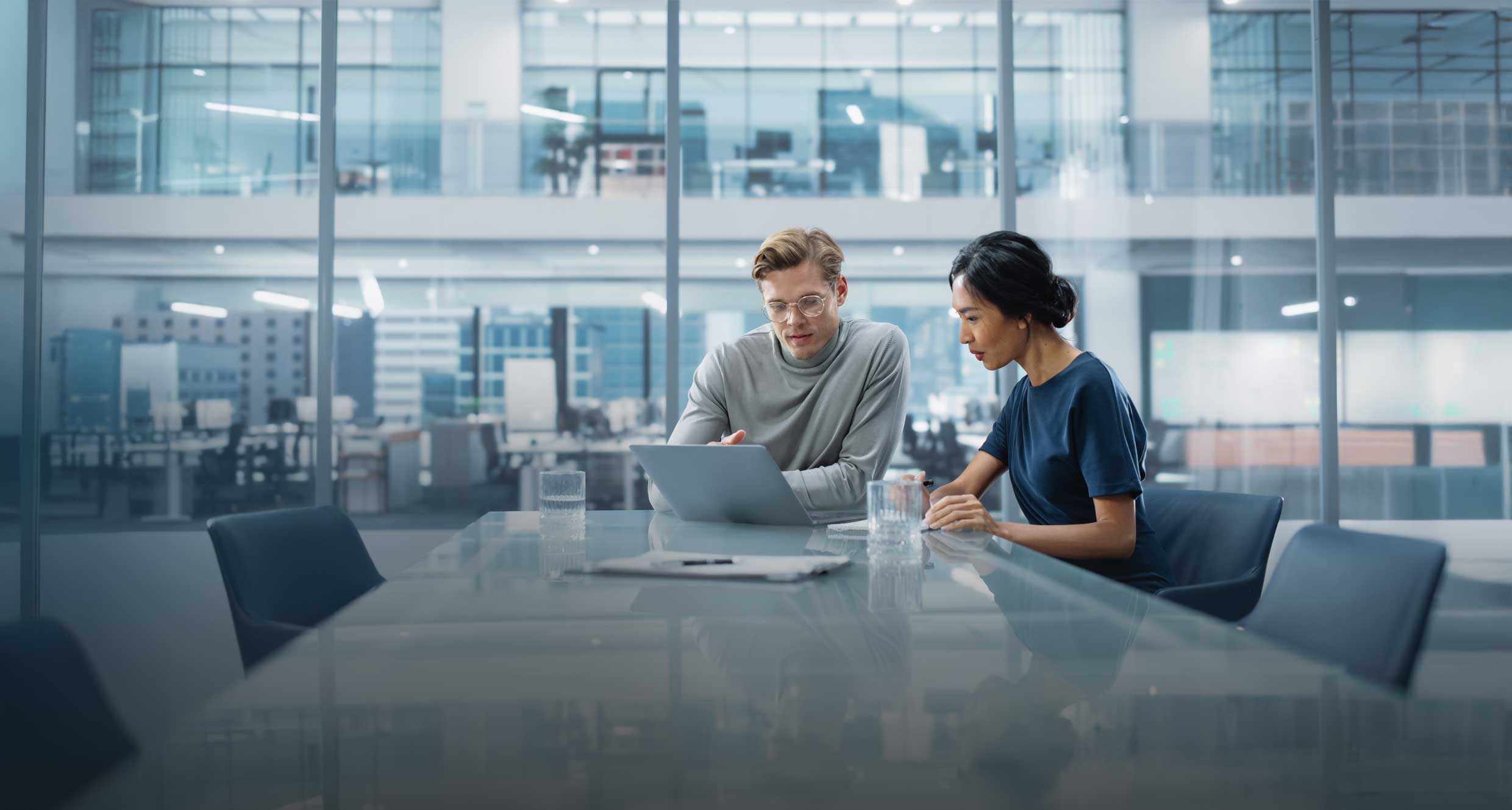 Two professional office workers sitting at a boardroom table, reviewing a laptop screen together.