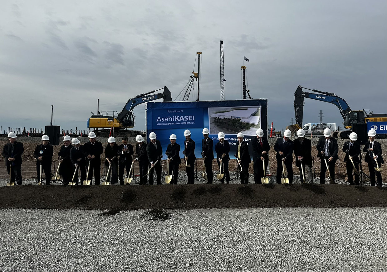Picture of men in suits and hardhats with shovels in front of Asahi Kasei sign with construction equipment